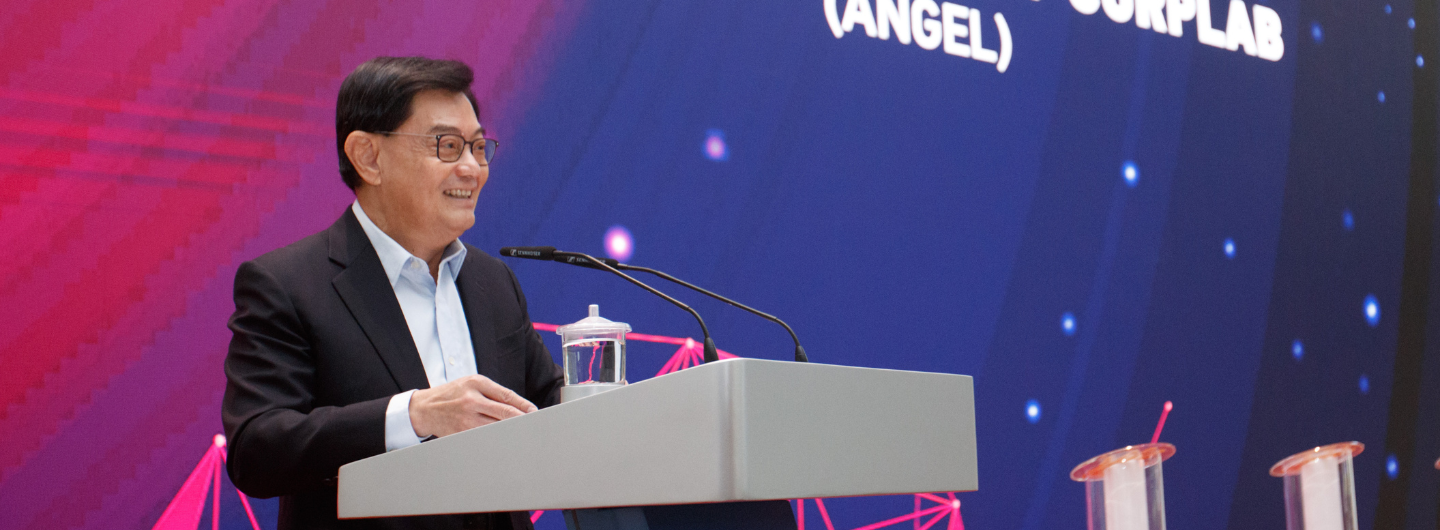 Man at podium with microphones, glass of water, blue screen reads "(ANGEL) SURPLAB."
